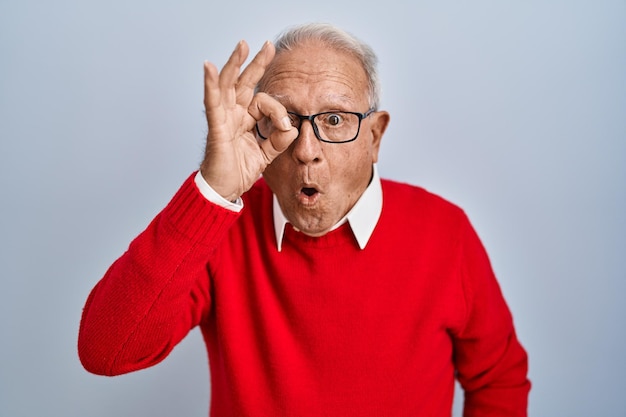 Homme senior aux cheveux gris debout sur fond isolé faisant un geste ok choqué par le visage surpris, les yeux regardant à travers les doigts. expression incrédule.