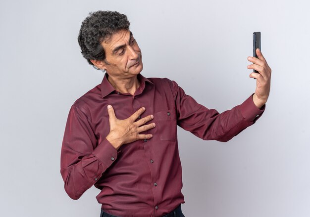 Homme senior en chemise violette faisant selfie à l'aide d'un smartphone à la confiance debout sur blanc