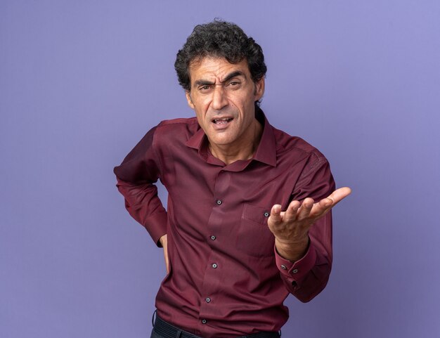 Homme senior en chemise violette regardant la caméra avec un visage en colère levant le bras pour se disputer ou poser une question