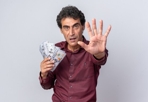 Homme senior en chemise violette tenant de l'argent en regardant la caméra avec un visage sérieux faisant un geste d'arrêt