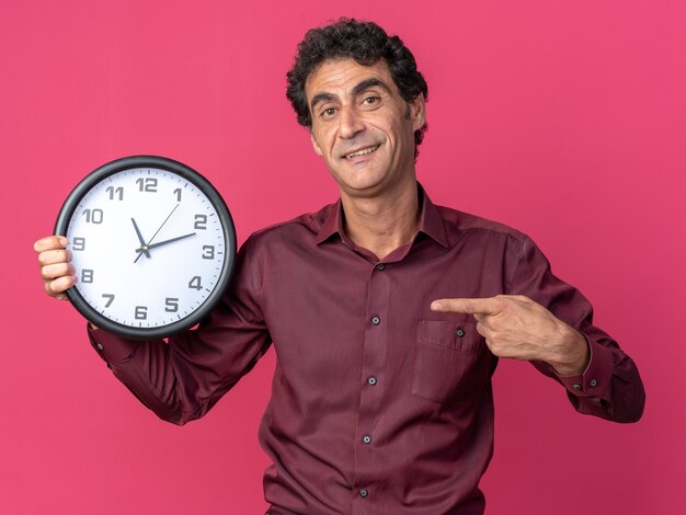 Homme senior en chemise violette tenant une horloge murale pointant avec l'index sur elle souriant heureux et positif debout sur fond rose