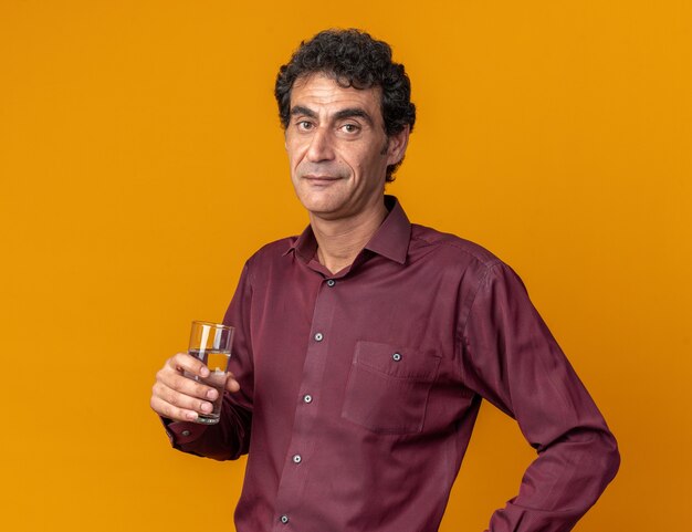 Homme senior en chemise violette tenant un verre d'eau regardant la caméra souriant confiant debout sur fond orange