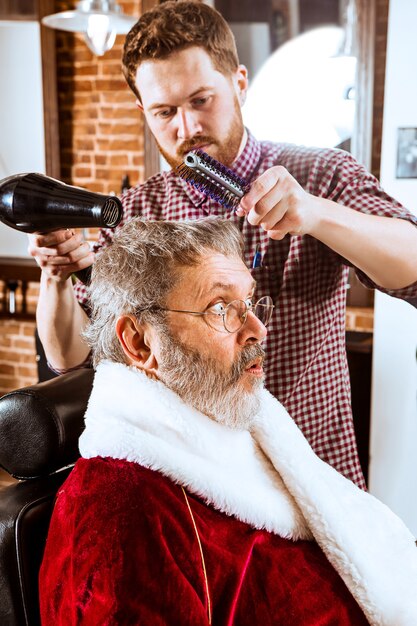 L'homme senior en costume de père Noël raser son maître personnel au salon de coiffure avant Noël