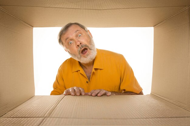 Homme senior étonné ouvrant le plus gros colis postal isolé sur blanc. Modèle masculin heureux sur le dessus de la boîte en carton regardant à l'intérieur.