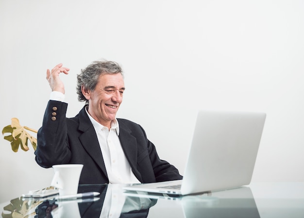 Homme senior excité souriant, assis sur le lieu de travail à la recherche d'un ordinateur portable