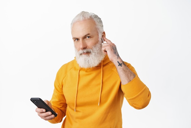 Homme senior hipster élégant mettre des écouteurs sans fil dans l'oreille tenant un smartphone et regardant la caméra va passer un appel téléphonique en écoutant de la musique dans des écouteurs portant un sweat à capuche fond blanc