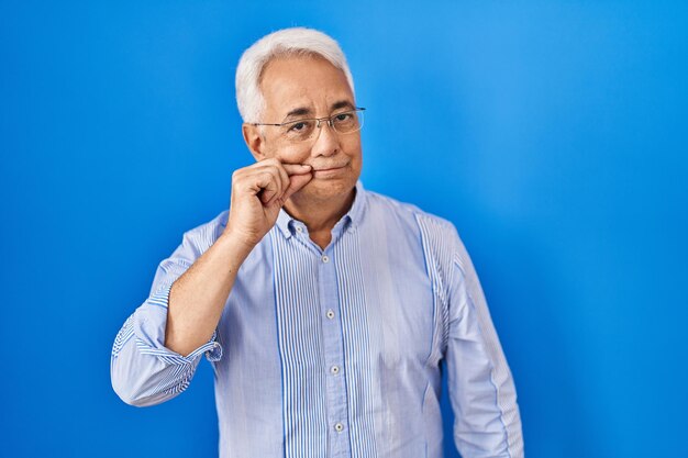 Homme senior hispanique portant des lunettes bouche et lèvres fermées comme zip avec les doigts. secret et silencieux, parler tabou