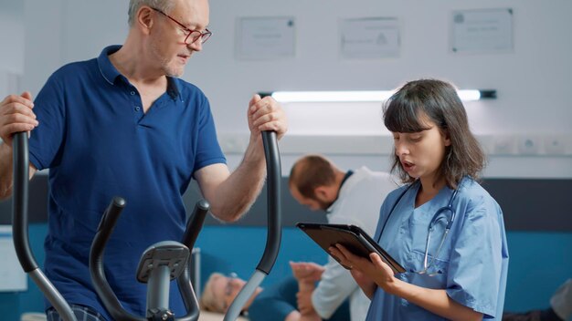 Homme senior et infirmière médicale parlant de la récupération de l'ostéopathie, patient utilisant un vélo électrique stationnaire pour augmenter la mobilité. Personne âgée assistant à une procédure de réadaptation pour guérir.