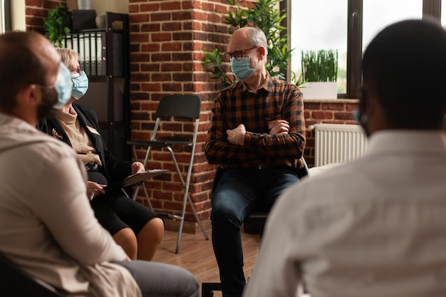 Homme senior avec masque facial parlant aux personnes en cercle lors d'une réunion, pour soigner la dépendance à l'alcool lors d'une thérapie de groupe de soutien. Personne ayant une discussion avec un psychologue sur des problèmes.