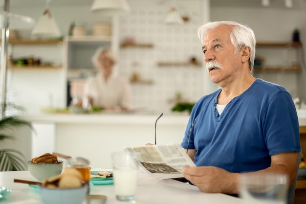 Homme senior pensif lisant le journal tout en prenant son petit déjeuner à la maison