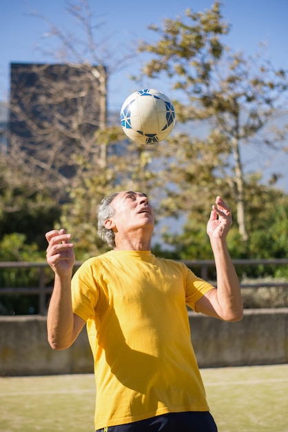 Photo gratuite homme senior satisfait avec ballon de football. homme de race blanche aux cheveux gris courts en vêtements de sport lançant une balle sur le terrain de sport seul. football, sport, concept de loisirs