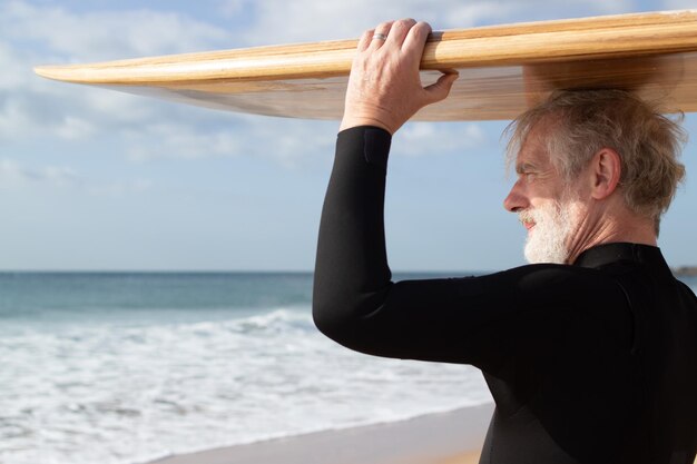 Homme senior sérieux tenant une planche de surf sur la tête. Homme barbu aux cheveux gris en combinaison avec planche de bois sur la tête regardant et profitant de la vue sur la mer et les vagues. Mode de vie actif et sain du concept de personnes âgées