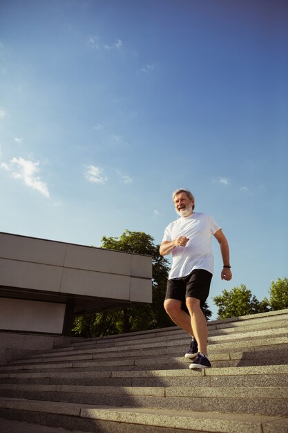 Homme senior en tant que coureur avec brassard ou tracker de fitness dans la rue de la ville. Modèle masculin caucasien pratiquant le jogging et les entraînements cardio le matin d'été. Mode de vie sain, sport, concept d'activité.