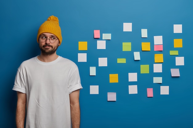 homme sérieux et contemplatif avec barbe, habillé avec désinvolture, pense à rédiger un diplôme, utilise des notes adhésives pour écrire des informations à retenir.