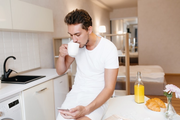 Homme sérieux avec coupe de cheveux courte vérifier le courrier sur le téléphone et boire du café