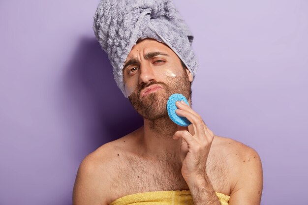 Homme sérieux détendu avec chaume essuie la peau du visage après la douche, tient une éponge cosmétique, enveloppée dans une serviette douce