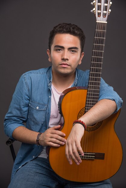 Homme sérieux avec guitare posant sur fond sombre.