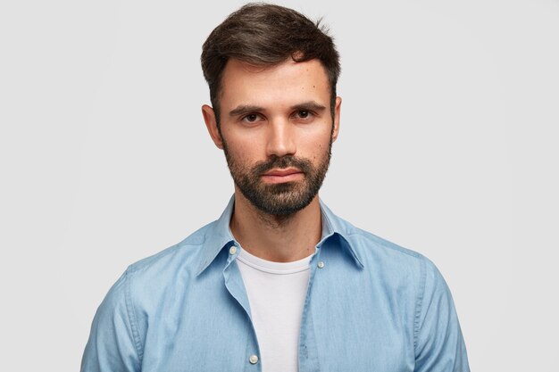 Homme sérieux mal rasé avec une apparence agréable, a les cheveux noirs, des poils, contemple quelque chose d'important, vêtu de vêtements à la mode, isolé sur un mur blanc. Homme européen à l'intérieur