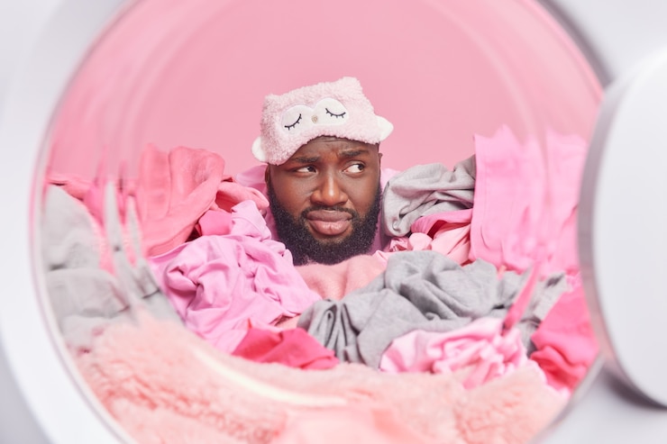 Photo gratuit un homme sérieux et réfléchi porte un masque de sommeil regarde au loin en étant plongé dans ses pensées tout en faisant des charges de linge machine à laver avec des vêtements sales pose contre le mur rose
