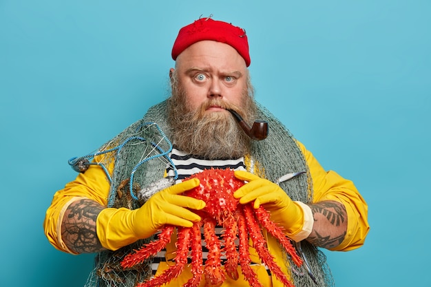 Homme sérieux strict avec une barbe épaisse, tient un gros crabe rouge, fume la pipe à tabac, aime la voile et la croisière, porte un chapeau rouge, un filet de pêche sur les épaules