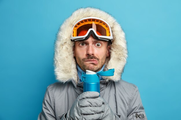 Photo gratuite un homme sérieux tremble de froid après avoir skié pendant une journée d'hiver glaciale tient un flacon avec une boisson chaude porte des lunettes de ski et une veste chaude.