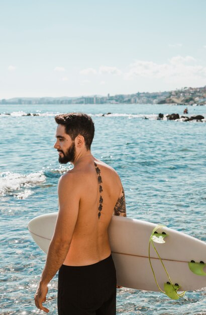 Homme en short debout avec une planche de surf blanche sur la plage