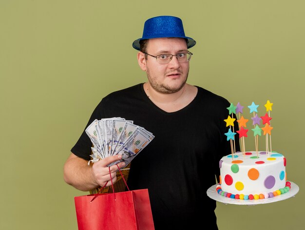 L'homme slave adulte anxieux dans des lunettes optiques portant un chapeau de fête bleu détient une boîte-cadeau d'argent sac à provisions en papier et gâteau d'anniversaire