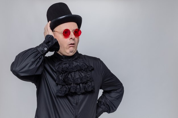 Homme slave adulte choqué avec chapeau haut de forme et lunettes de soleil en chemise gothique noire regardant de côté