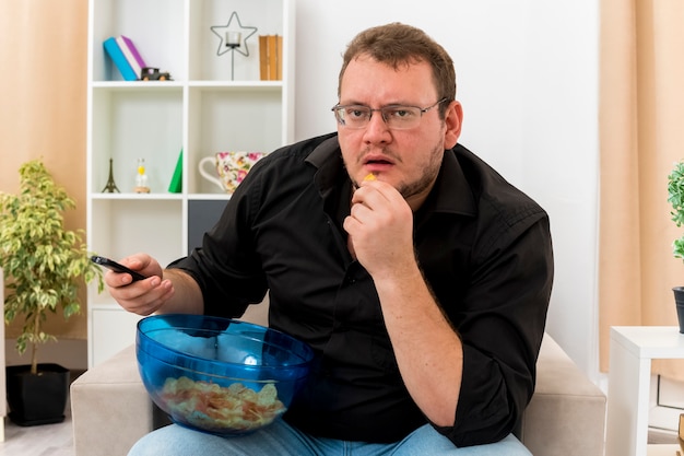 Homme slave adulte choqué dans des lunettes optiques est assis sur un fauteuil tenant la télécommande du téléviseur et un bol de chips sur les jambes à l'intérieur du salon