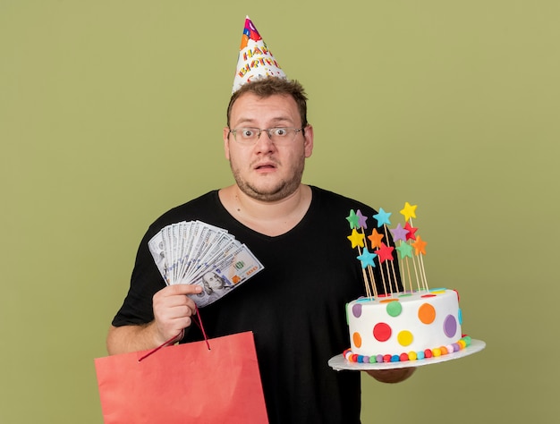 Un homme slave adulte choqué dans des lunettes optiques portant une casquette d'anniversaire tient un sac à provisions en papier d'argent et un gâteau d'anniversaire