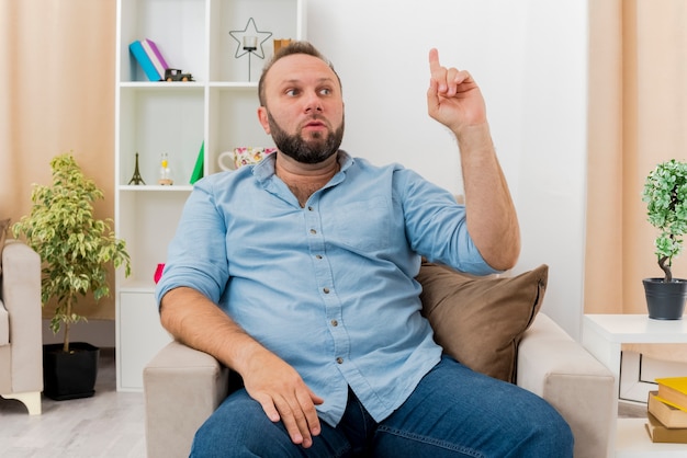 Homme slave adulte choqué est assis sur un fauteuil pointant vers le haut et regardant sur le côté à l'intérieur du salon