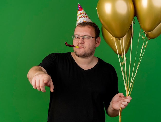 Un homme slave adulte confiant dans des lunettes optiques portant une casquette d'anniversaire tient des ballons à l'hélium soufflant un sifflet de fête et pointant vers la caméra