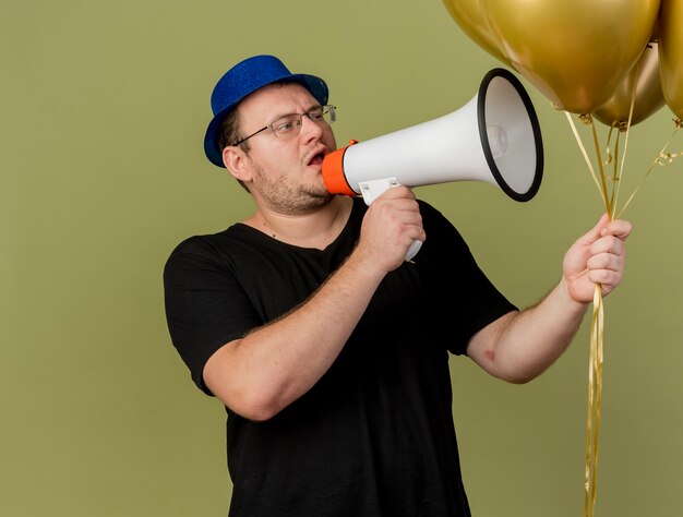 Un homme slave adulte confiant dans des lunettes optiques portant un chapeau de fête bleu tient et regarde des ballons à l'hélium parlant dans un haut-parleur