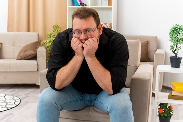 Homme slave adulte déçu dans des lunettes optiques est assis sur un fauteuil met les mains sur le menton à l'intérieur de la salle de séjour conçue