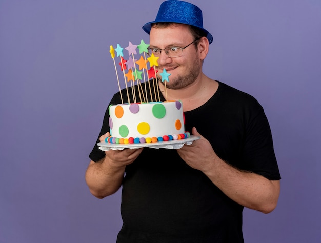 Homme slave adulte excité dans des lunettes optiques portant un chapeau de fête bleu tient et regarde le gâteau d'anniversaire