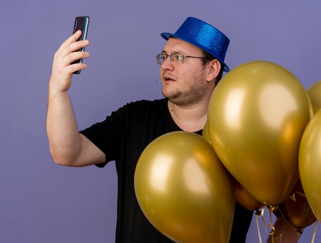 Un homme slave adulte impressionné dans des lunettes optiques portant un chapeau de fête bleu se tient avec des ballons à l'hélium en regardant le téléphone