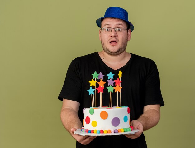 Un homme slave adulte impressionné dans des lunettes optiques portant un chapeau de fête bleu tient un gâteau d'anniversaire