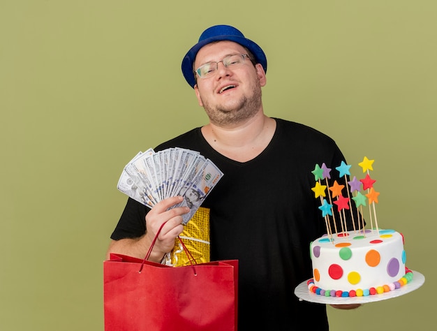 Un homme slave adulte joyeux dans des lunettes optiques portant un chapeau de fête bleu contient un sac à provisions en papier et un gâteau d'anniversaire