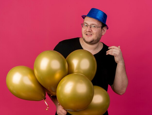 Un homme slave adulte joyeux dans des lunettes optiques portant un chapeau de fête bleu tient des ballons à l'hélium