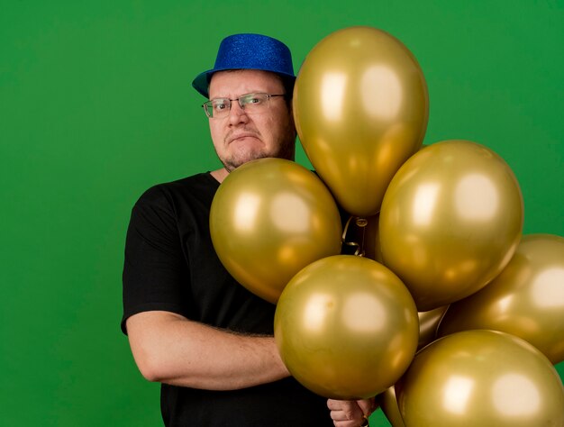 Un homme slave adulte mécontent dans des lunettes optiques portant un chapeau de fête bleu tient des ballons à l'hélium