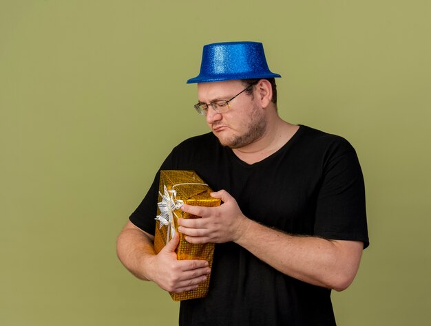 Homme slave adulte mécontent dans des lunettes optiques portant un chapeau de fête bleu tient et regarde la boîte-cadeau
