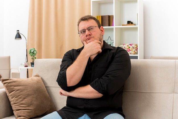 Homme slave adulte réfléchi dans des lunettes optiques est assis sur un fauteuil tenant le menton et regardant sur le côté à l'intérieur de la salle de séjour