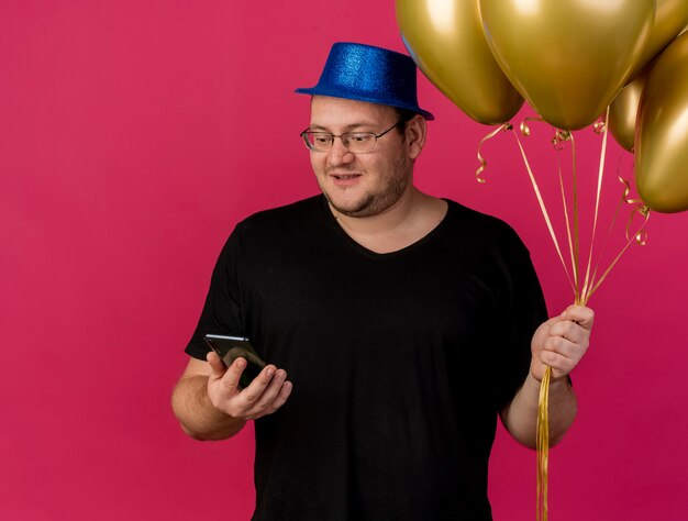 Un homme slave adulte surpris dans des lunettes optiques portant un chapeau de fête bleu tient des ballons à l'hélium et regarde le téléphone