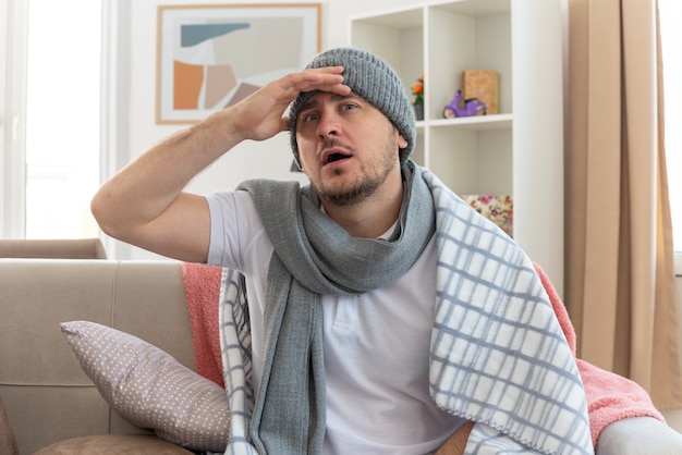 Homme slave malade surpris avec une écharpe autour du cou portant un chapeau d'hiver enveloppé dans un plaid mettant la main sur le front assis sur un canapé dans le salon