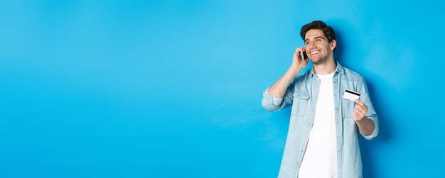 Un homme souriant appelle le support bancaire en parlant sur un téléphone portable et en tenant une carte de crédit debout contre le bleu