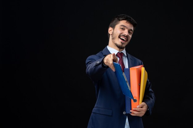 Homme souriant en costume tenant plusieurs documents et pointant vers l'avant sur un mur sombre isolé