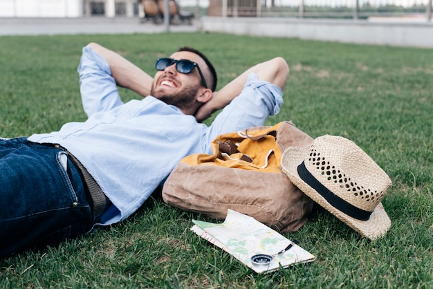 Homme souriant détendu, couché sur l'herbe avec accessoires de voyage