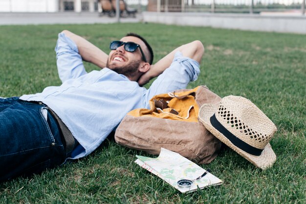 Homme souriant détendu, couché sur l'herbe avec accessoires de voyage