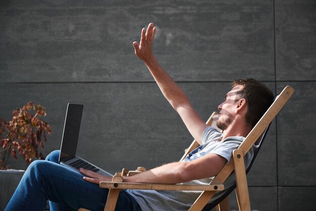 Un homme souriant est assis sur une chaise avec un ordinateur portable en plein air