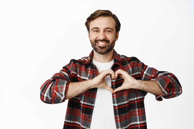 Homme souriant montrant signe de coeur debout en chemise à carreaux sur fond blanc Espace de copie